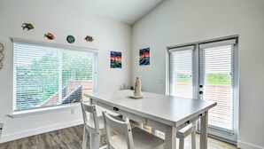 Un coin repas avec une table et des chaises blanches, une fenêtre avec des stores et des œuvres d’art murales.