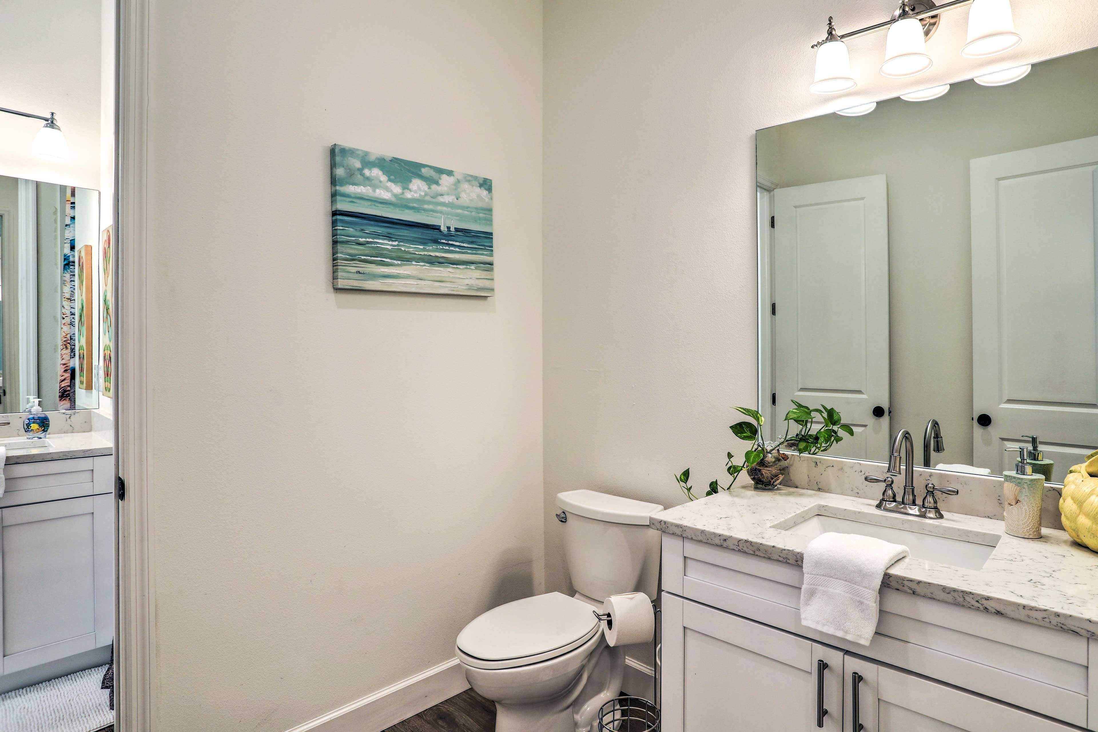 Un baño con inodoro, lavabo y espejo. Hay un cuadro en la pared y una planta en la encimera.