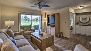 A living room with a sofa, coffee table, and TV. There's a ceiling fan, a window with a view, and a kitchen area with cabinets and a microwave. - St Pete Condo w/ Heated Pool - 3 Miles to Beach! (St. Petersburg)