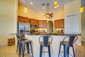 A kitchen with wooden cabinets, a stainless steel refrigerator, a microwave, a ceiling fan, and three bar stools. - 3 Mi to Downtown Cape Coral: Canal-front Villa (Cape Coral)