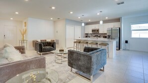 Uma sala de estar espaçosa com um sofá cinzento, uma mesa de centro de vidro e uma cozinha moderna com armários brancos e eletrodomésticos de aço inoxidável.