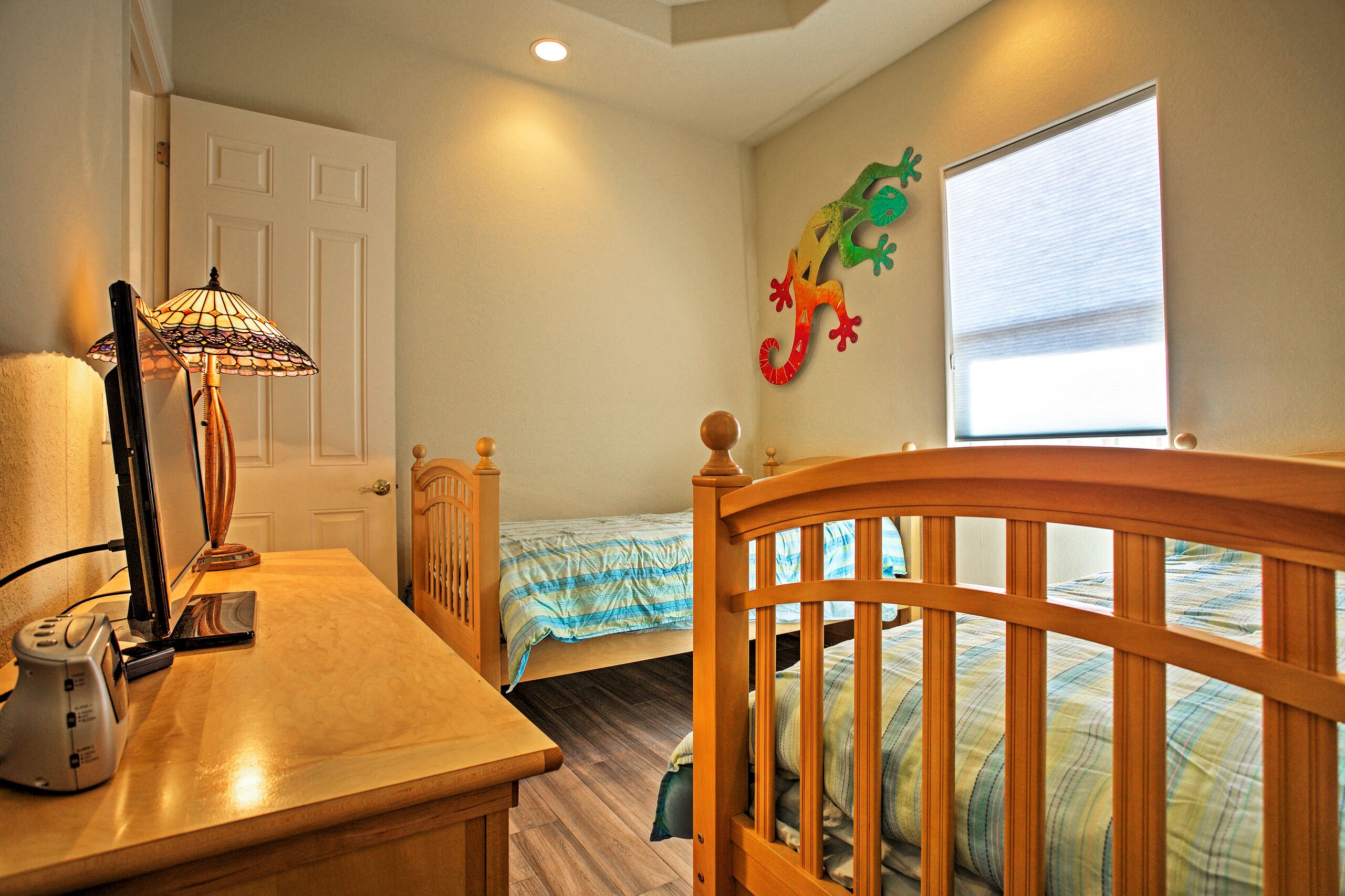 A bedroom with a wooden bed, a desk with a lamp, a window with blinds, and a wall decoration of a lizard.