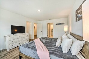 Quarto com cama, cômoda, televisão e corredor que dá acesso a outros espaços.