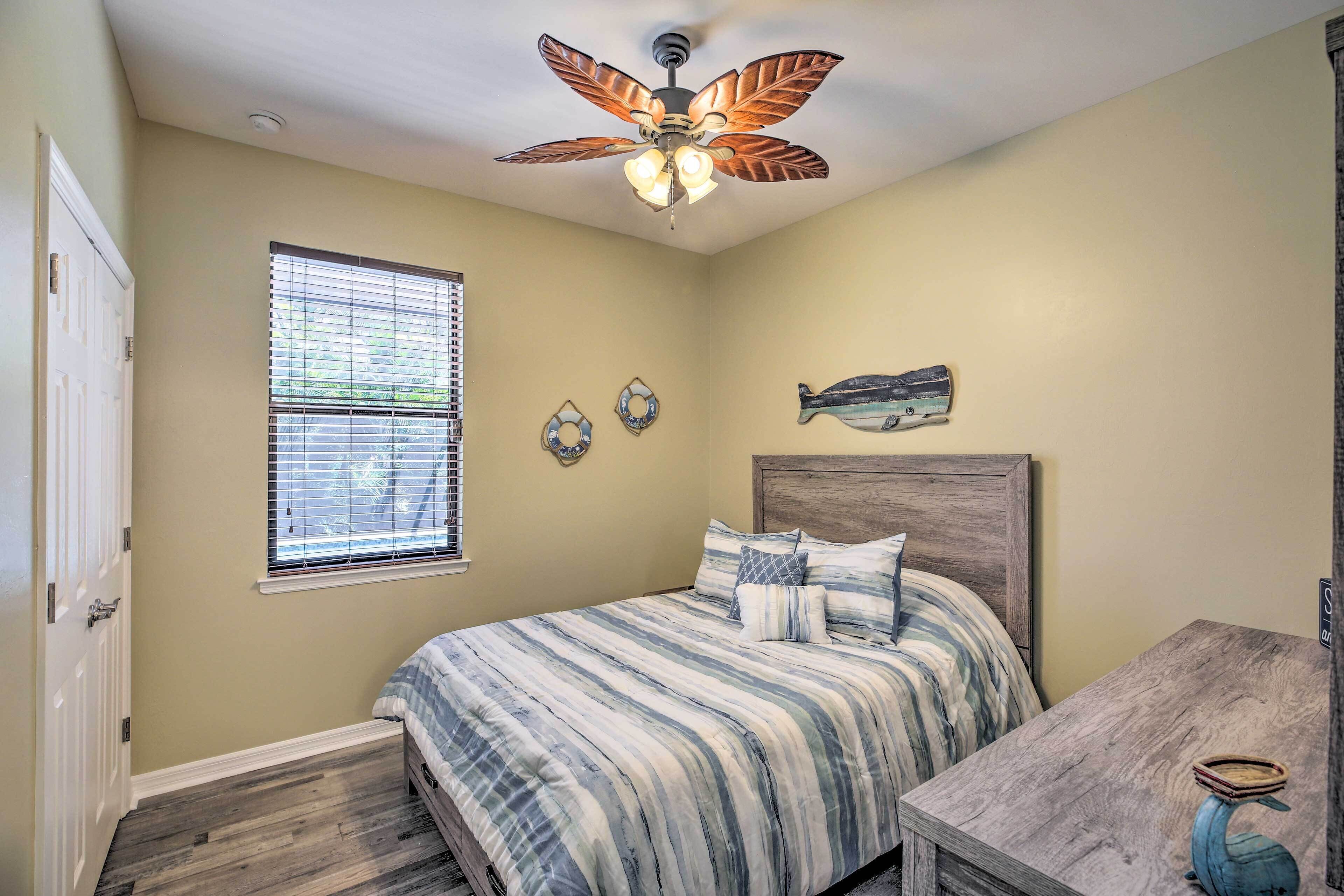 A bedroom with a bed, a ceiling fan, a window with blinds, and a small table with a decorative item.