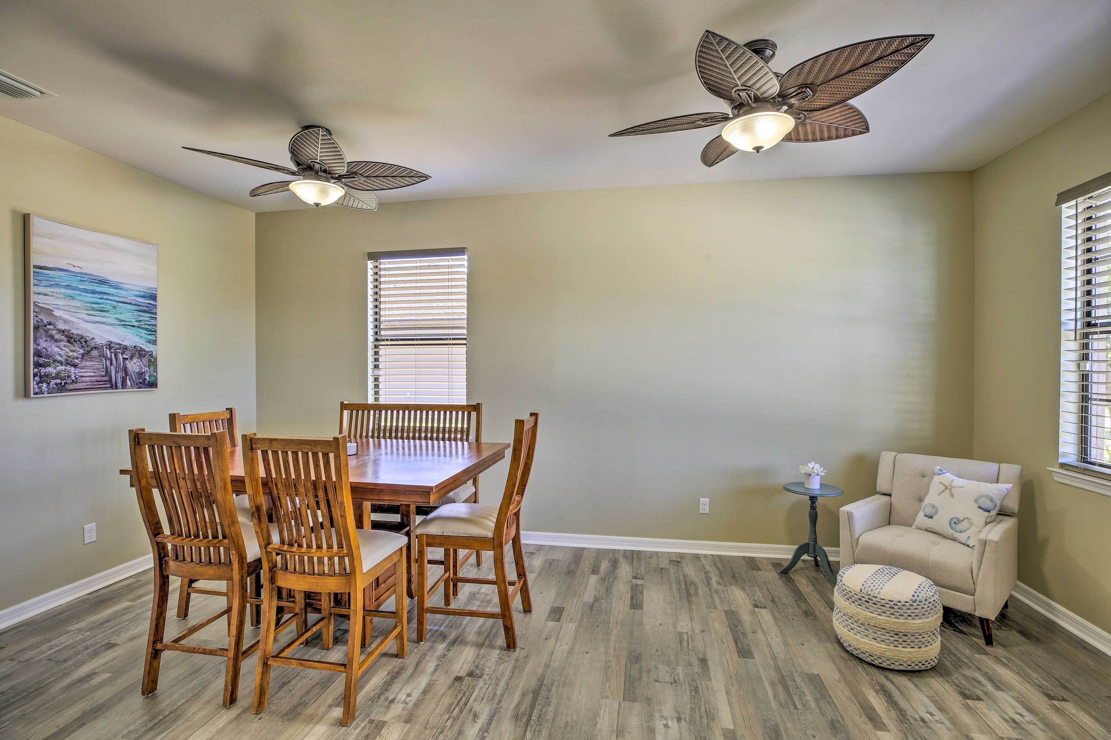 A dining area with a wooden table and chairs, two ceiling fans, a window with blinds, a small round table with a lamp, and a cushioned armchair.