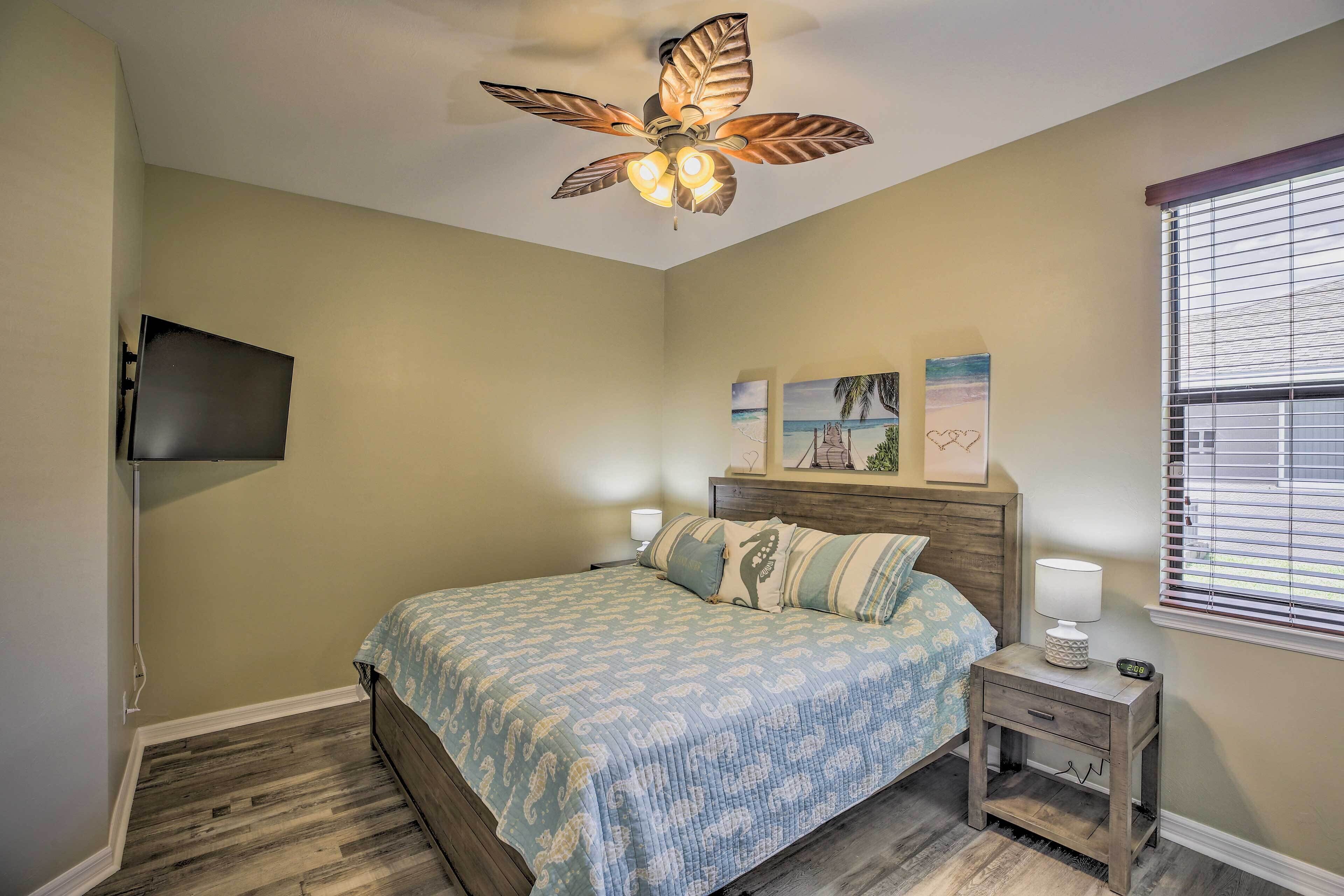 A bedroom with a bed, a nightstand, a ceiling fan, a TV mounted on the wall, and a window with blinds.