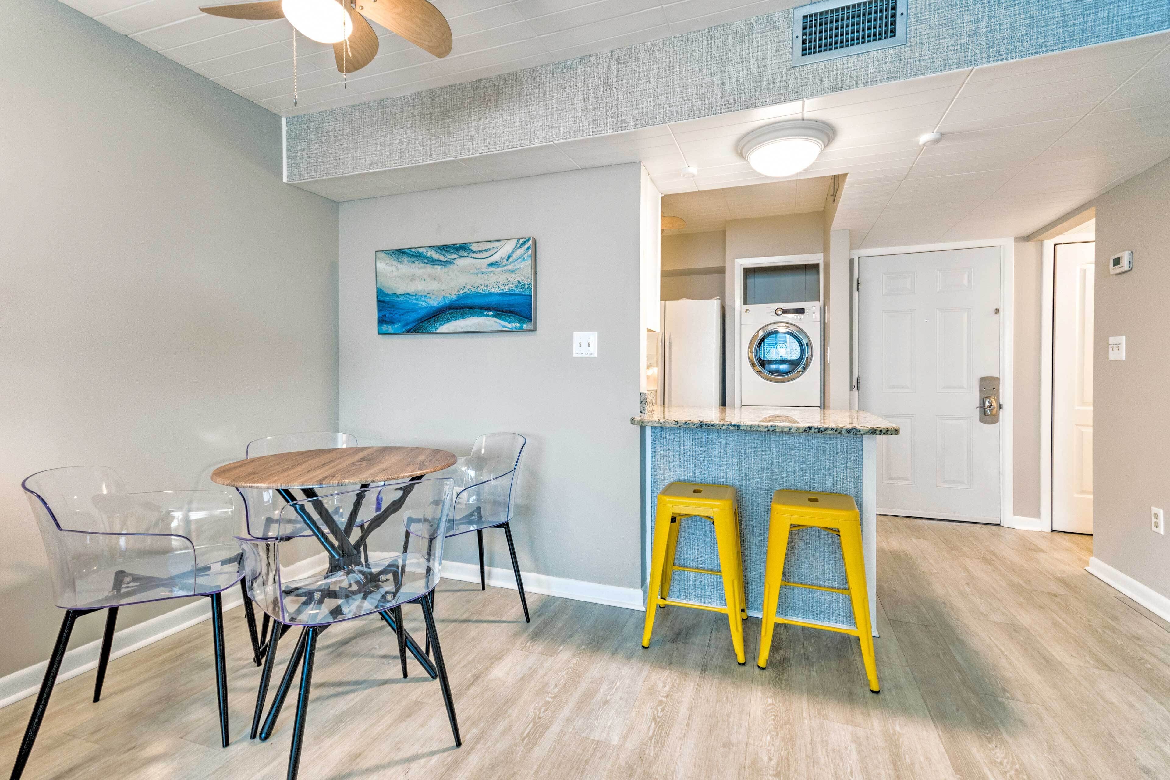 A modern kitchen with a built-in washer and dryer, a dining area with a round table and clear chairs, and a ceiling fan.