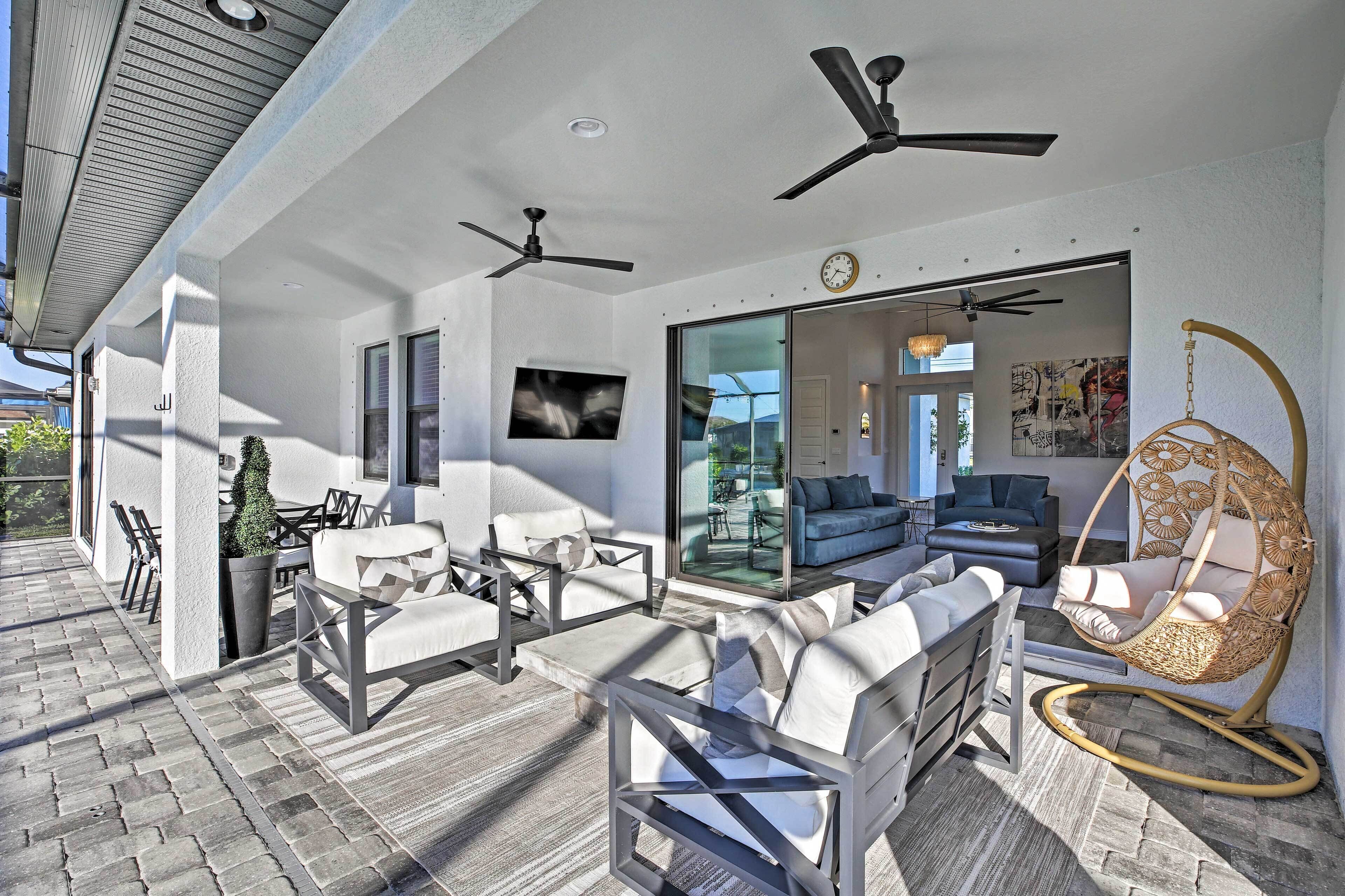 Un espacio exterior moderno con mobiliario, ventiladores de techo y un amplio ventanal que da a un ambiente interior.