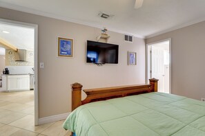 A bedroom with a bed, a television on the wall, and a kitchen visible through an open door.