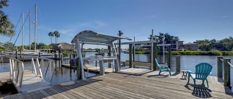 Un muelle de madera con zona de estar cubierta y sillas, con vista a una masa de agua tranquila, con barcos y palmeras al fondo.