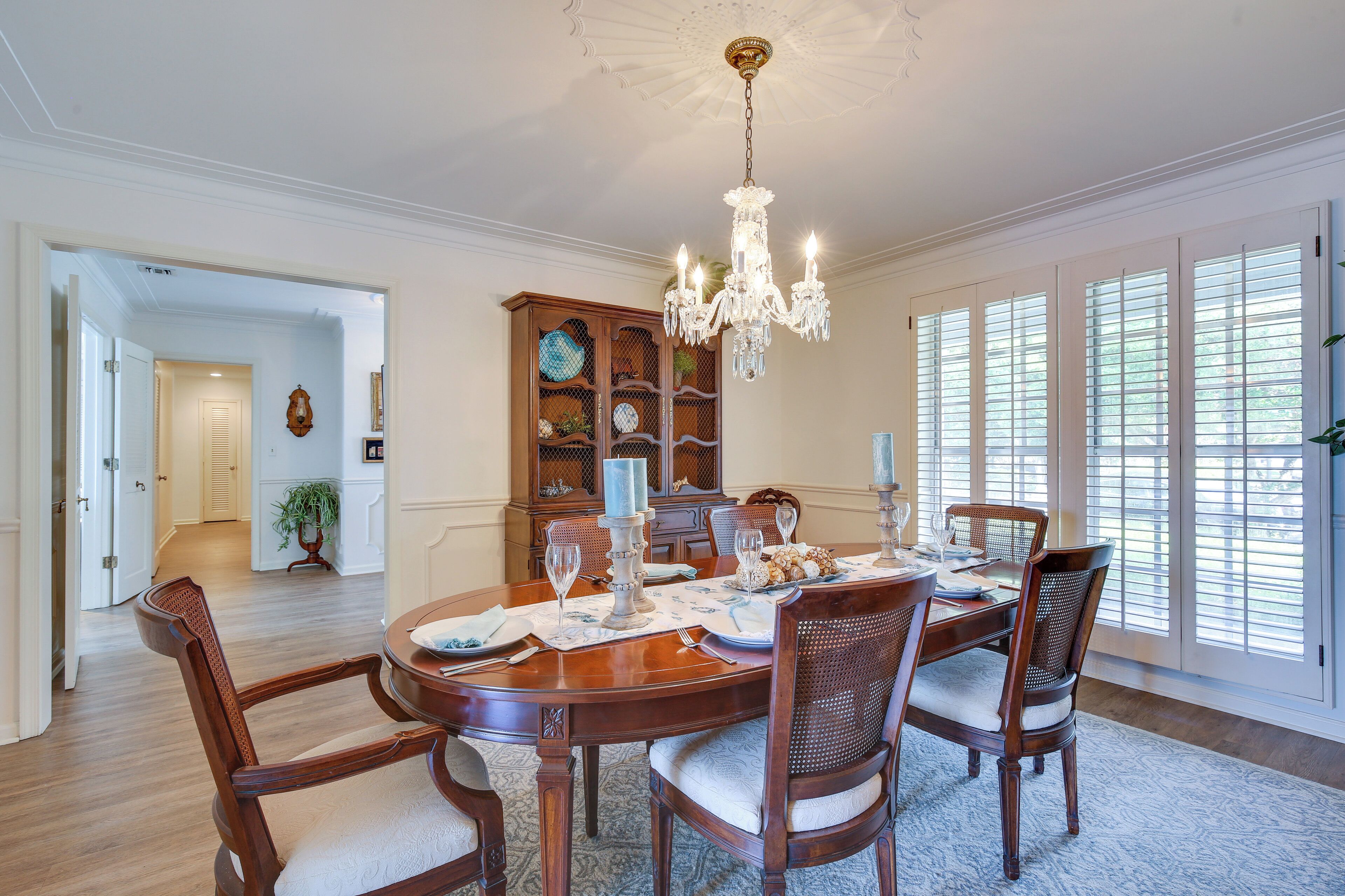 A dining room with a wooden table set for six, surrounded by six chairs, a chandelier, and a large window with blinds.