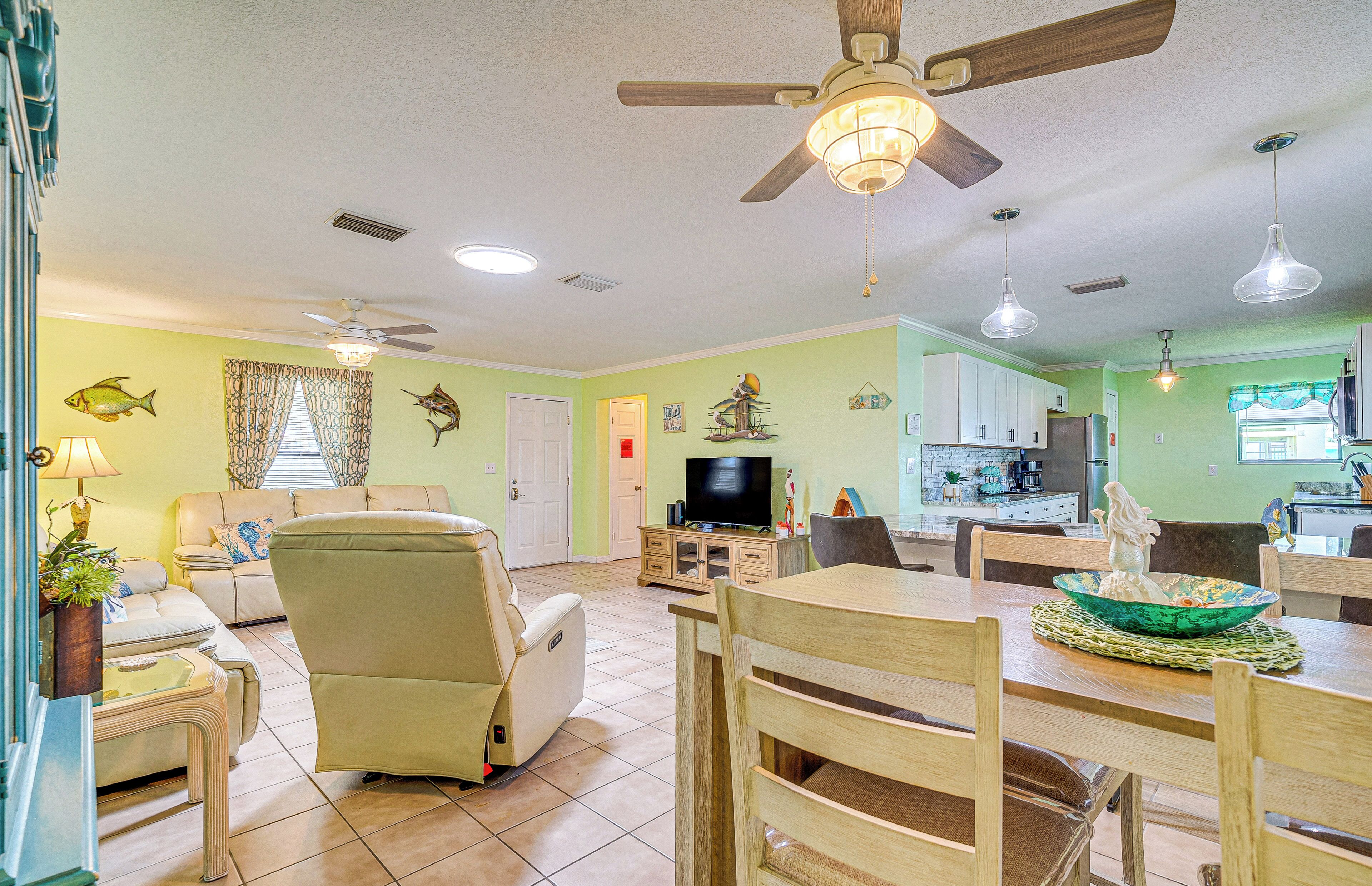 A spacious living room with a sofa, a recliner, a dining table with chairs, a ceiling fan, and a kitchen area with cabinets and a window.