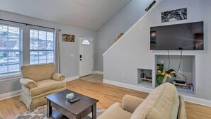 Ein Wohnzimmer mit einer Couch, einem Sessel und einem Couchtisch. Ein Fernseher ist an der Wand montiert, und es gibt eine Tür zu einem anderen Raum.