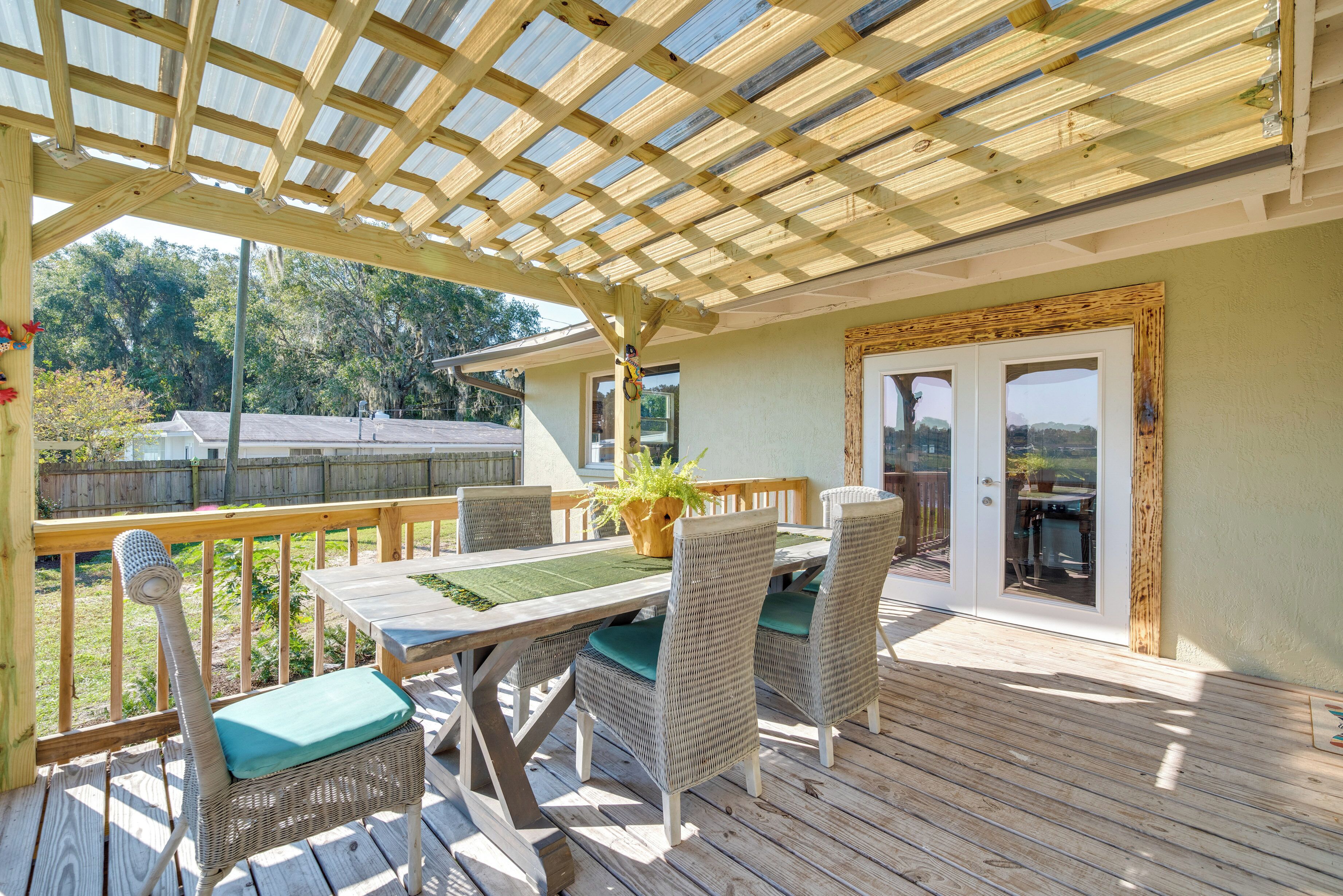 Une terrasse extérieure couverte, avec une table en bois et des chaises en osier, une pergola au-dessus et une vue sur une maison en arrière-plan.