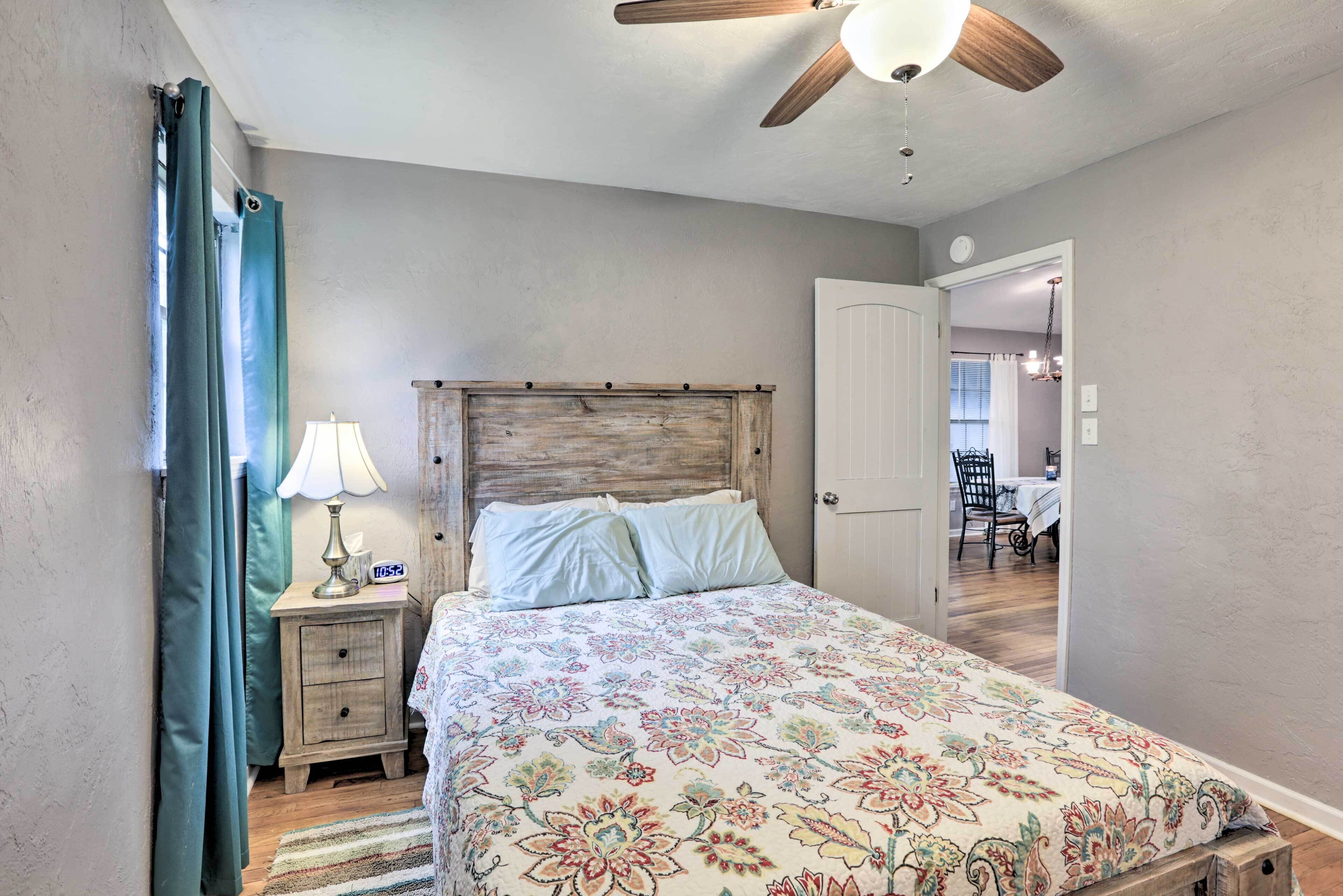 A bedroom with a wooden headboard, a floral bedspread, a nightstand with a lamp, and a view into the dining area.