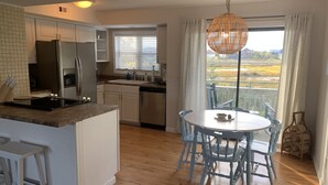 A modern kitchen with white cabinets, a built-in oven, and a stainless steel refrigerator. There is a dining area with a round table and blue chairs, and a large window offering a view of a natural landscape. - Carolina Marsh Hideaway (Carolina Beach)