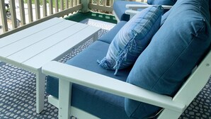 A covered outdoor seating area with a white bench and blue cushions, overlooking a residential street with houses and palm trees. - Carolina Marsh Hideaway (Carolina Beach)