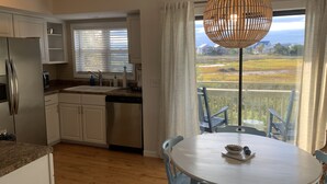 A kitchen with white cabinets, a stainless steel refrigerator, and a dishwasher. There's a window with blinds, a dining table with chairs, and a view of a landscape through the window. - Carolina Marsh Hideaway (Carolina Beach)