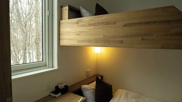 A compact hotel room with a bunk bed, a bedside table, a lamp, and a window with a view of trees.