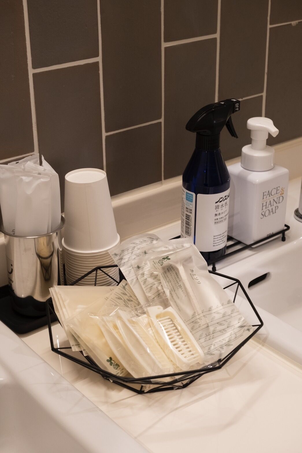Uma bancada de banheiro com um dosador de sabonete líquido, um borrifador e uma pilha de copos brancos.