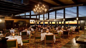 Un restaurant doté de nombreuses tables et chaises, d’un lustre et de grandes fenêtres donnant sur un coucher de soleil.