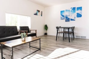 A modern living room with a sofa, coffee table, dining table, chairs, and artwork on the walls.