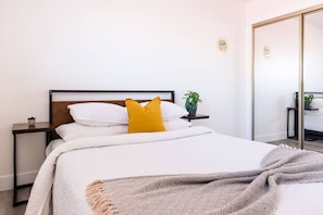 A neatly made bed with white bedding and a single yellow pillow, a bedside table with a plant, and a mirror on the wall.