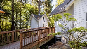 Une terrasse en bois mène à une cabane blanche dotée d’une cheminée, le tout étant entouré d’arbres.