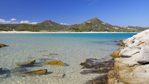 Una playa costera con aguas turquesas cristalinas, costa rocosa y una montaña al fondo.