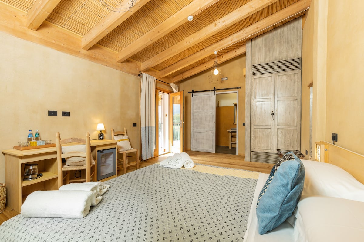 A hotel room with a large bed, wooden ceiling beams, a desk with a chair, a television, and a wardrobe.