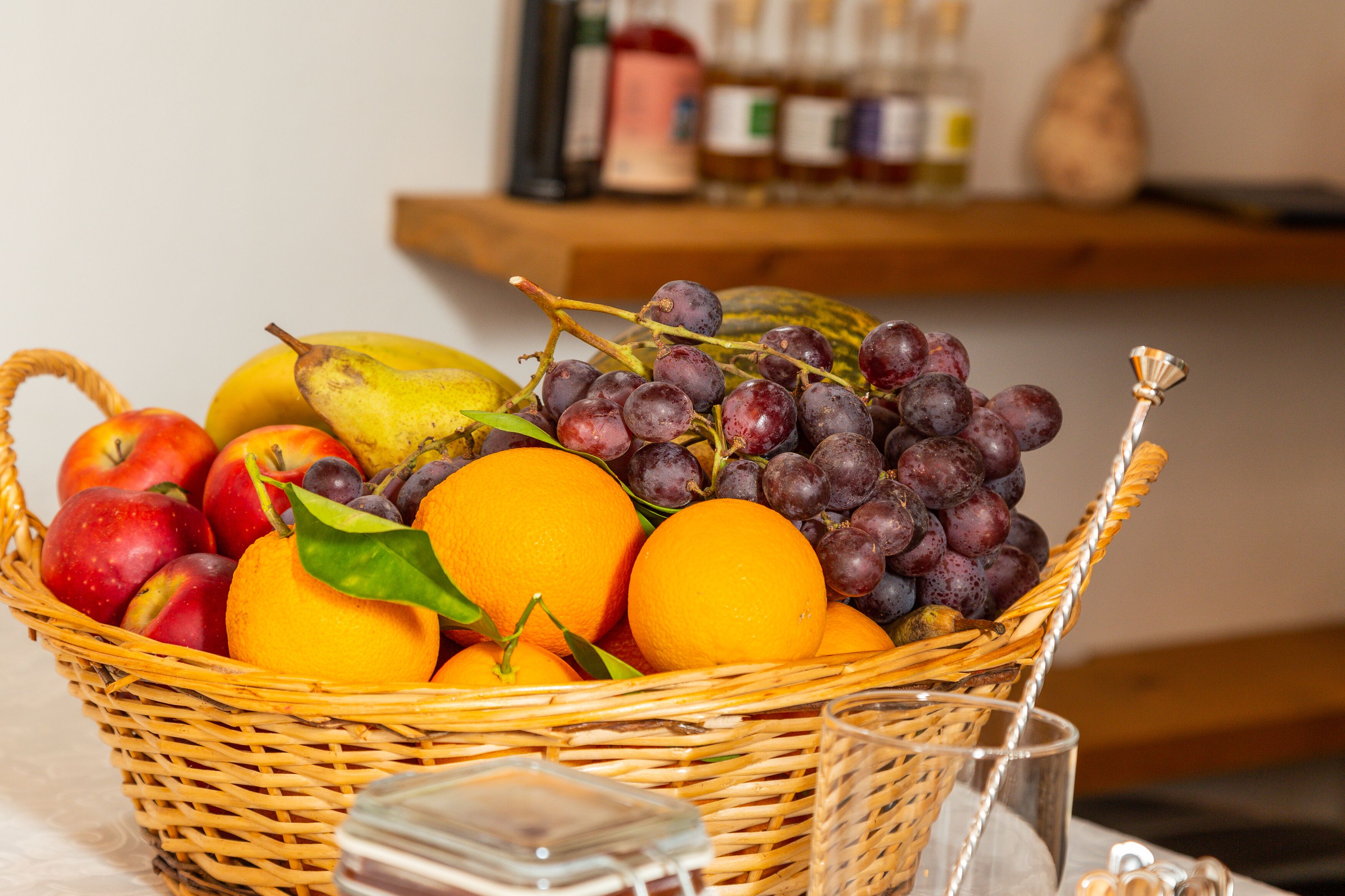 A wicker basket filled with fruits including apples, oranges, grapes, and a pear, placed on a table with a glass and a small container.