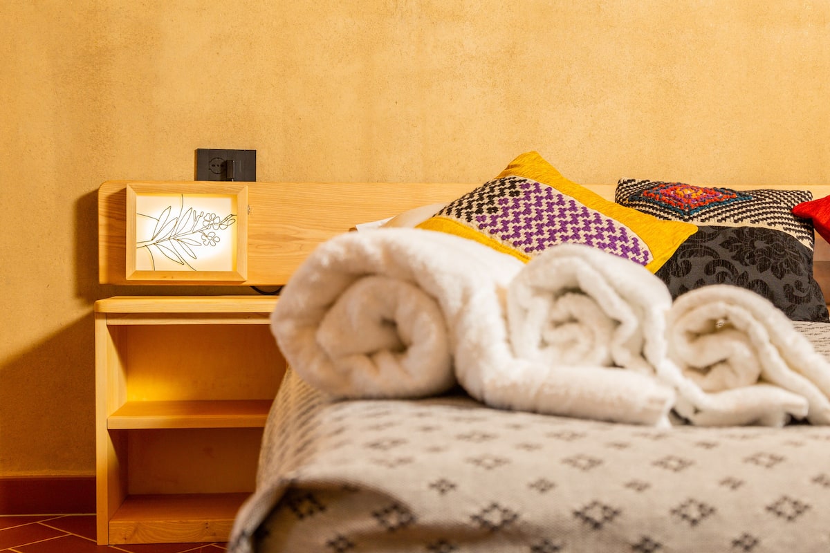 A neatly made bed with rolled white towels, a framed artwork on the headboard, and a wooden nightstand with a power outlet.