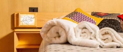 A neatly made bed with rolled white towels, a framed artwork on the headboard, and a wooden nightstand with a power outlet.