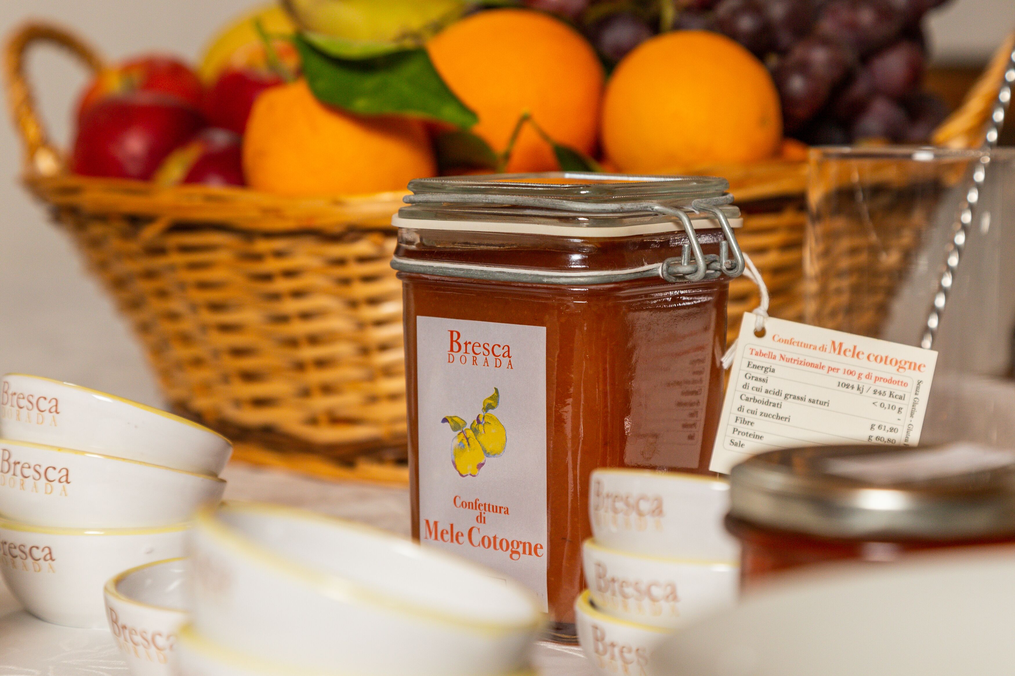 Un tarro de miel Bresca Dorada con etiqueta, rodeado de fruta y juegos de cucharillas para miel.