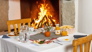 Una mesa de comedor dispuesta con platos, una jofaina, una vela y un cuenco de pan, con una chimenea al fondo.