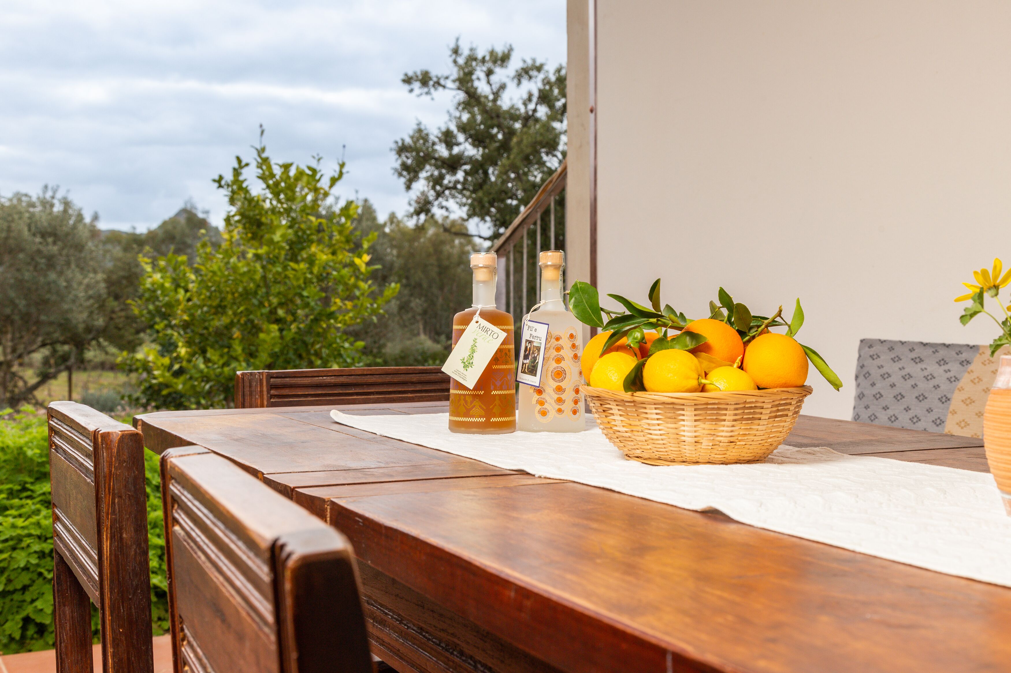 A wooden table with a white tablecloth, two bottles, and a basket of lemons and oranges on an outdoor patio with a view of trees and sky.