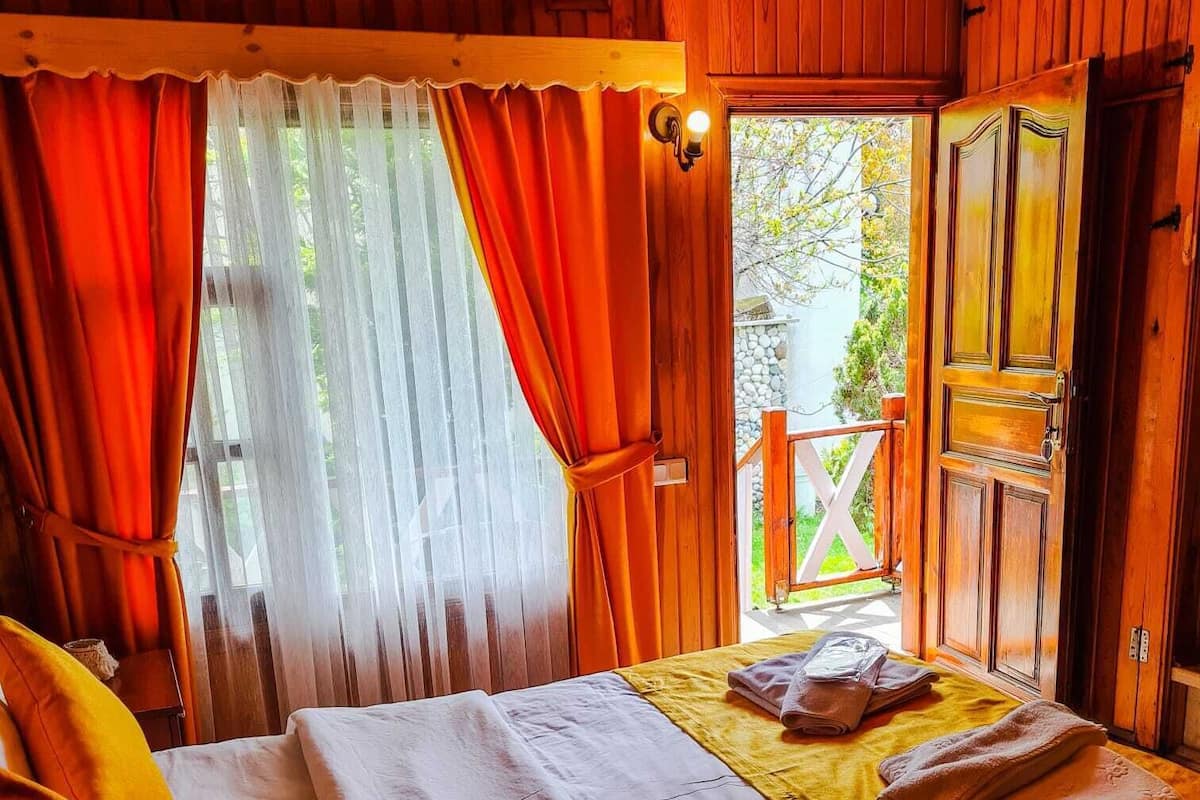 A wooden cabin bedroom with a bed, curtains, and a view of the outside.