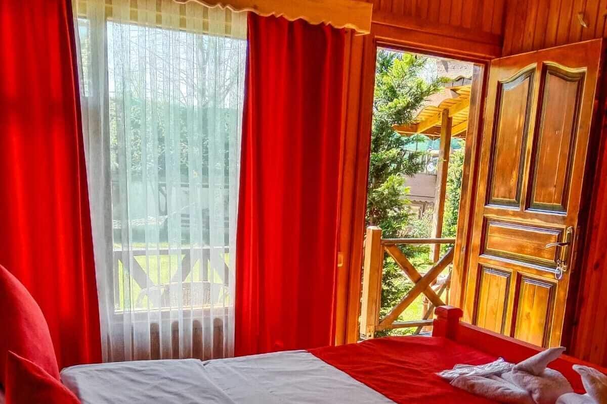 A room with a bed, red curtains, and a view of a wooden deck and greenery outside.