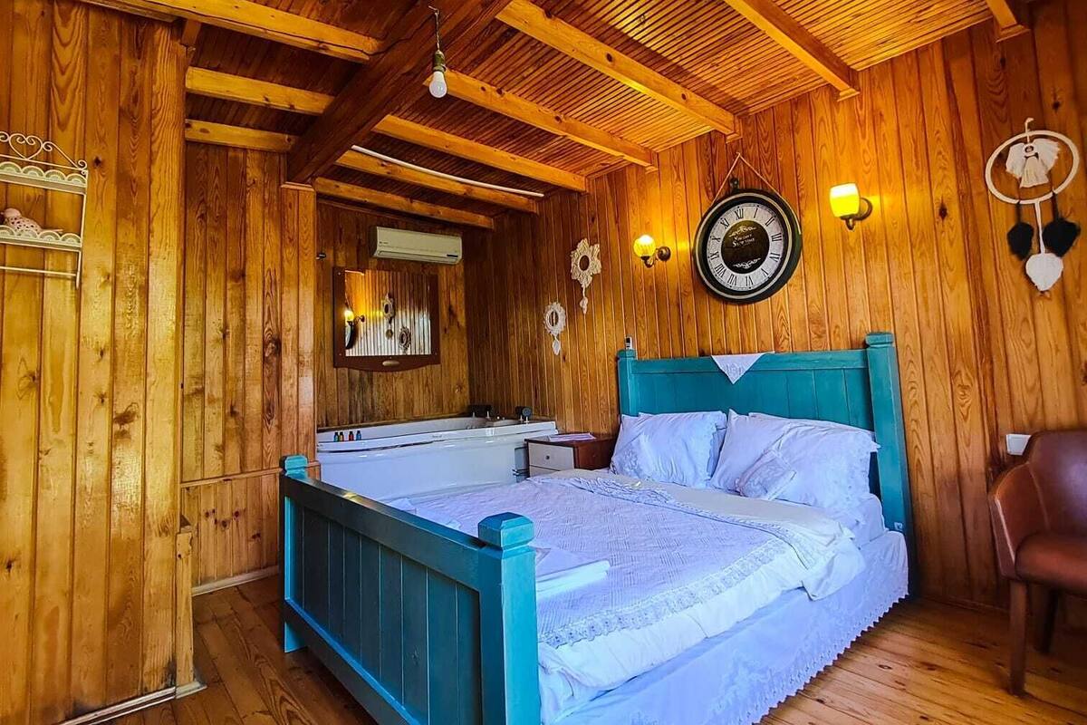 A wooden cabin bedroom with a bed, a wall clock, and a wall-mounted light fixture.