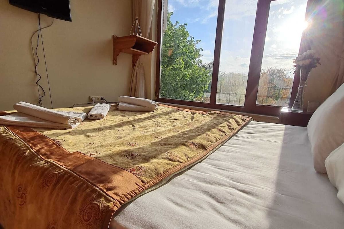 A hotel room with a large bed, a flat-screen TV mounted on the wall, a window with a view of trees, and a small wooden shelf.