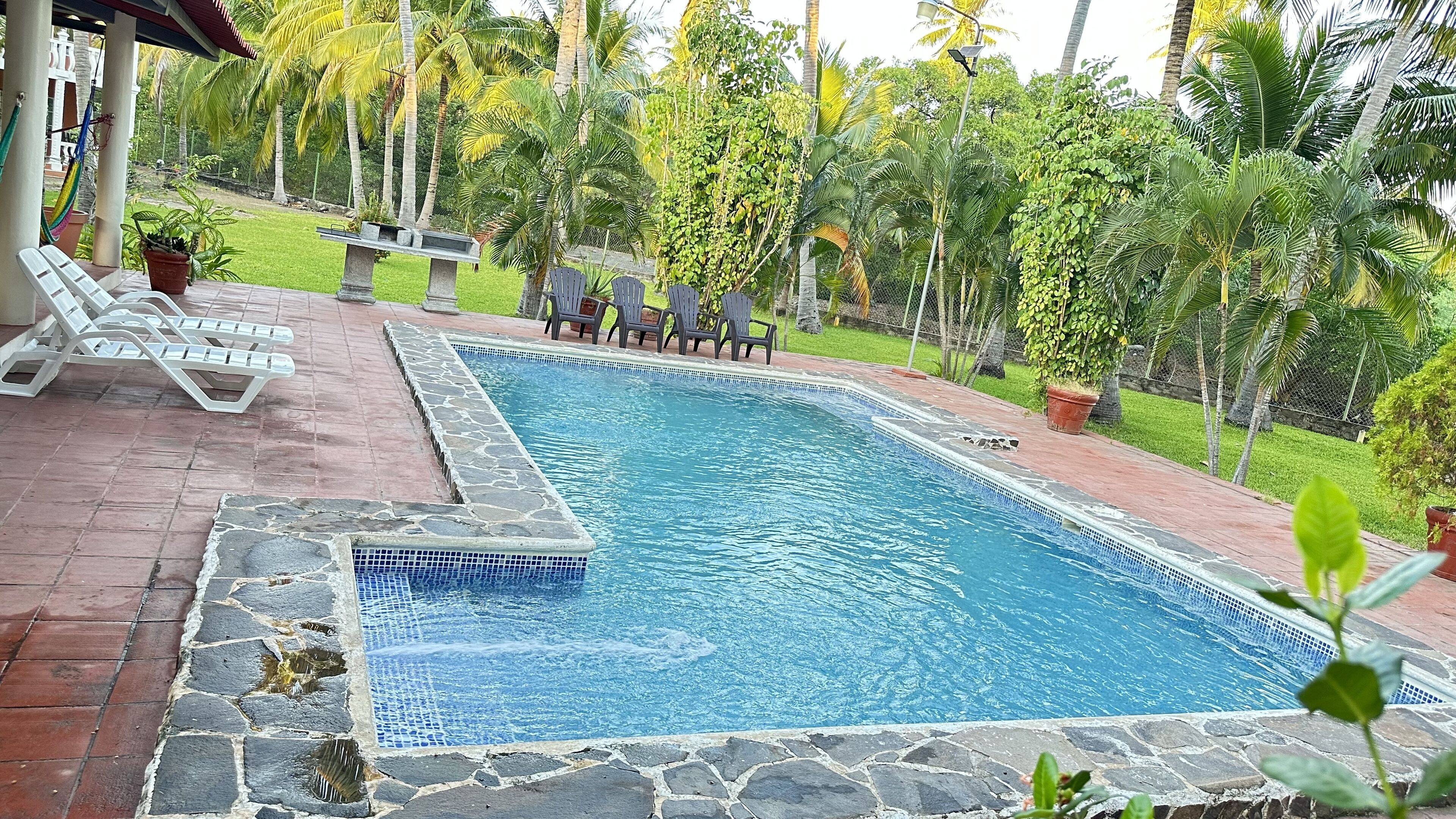 Ein rechteckiger Pool mit Steinfliesen, umgeben von einer Ziegelterrasse. Auf der Terrasse stehen Liegestühle und ein kleiner Tisch.