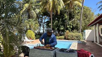 A man grilling outdoors with a pool and palm trees in the background.