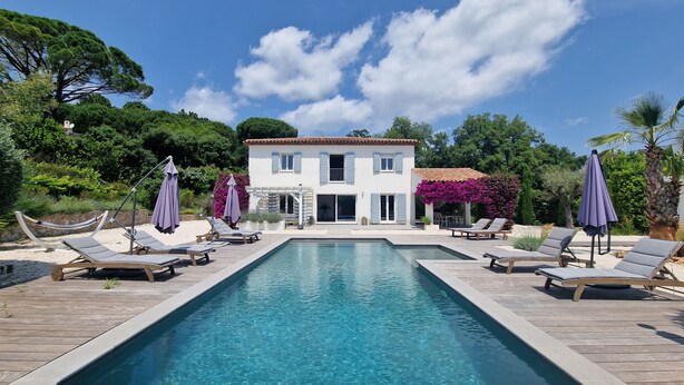 Un espace aménagé au bord de la piscine, avec des chaises longues et des parasols, une piscine et une maison à deux étages avec un toit en tuiles.