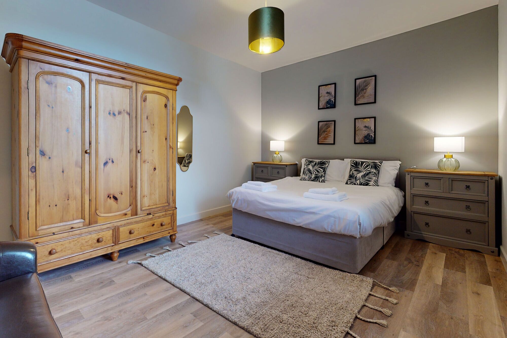 A bedroom with a large wooden wardrobe, a bed with white linens, two bedside tables with lamps, and a grey wall with framed artwork.