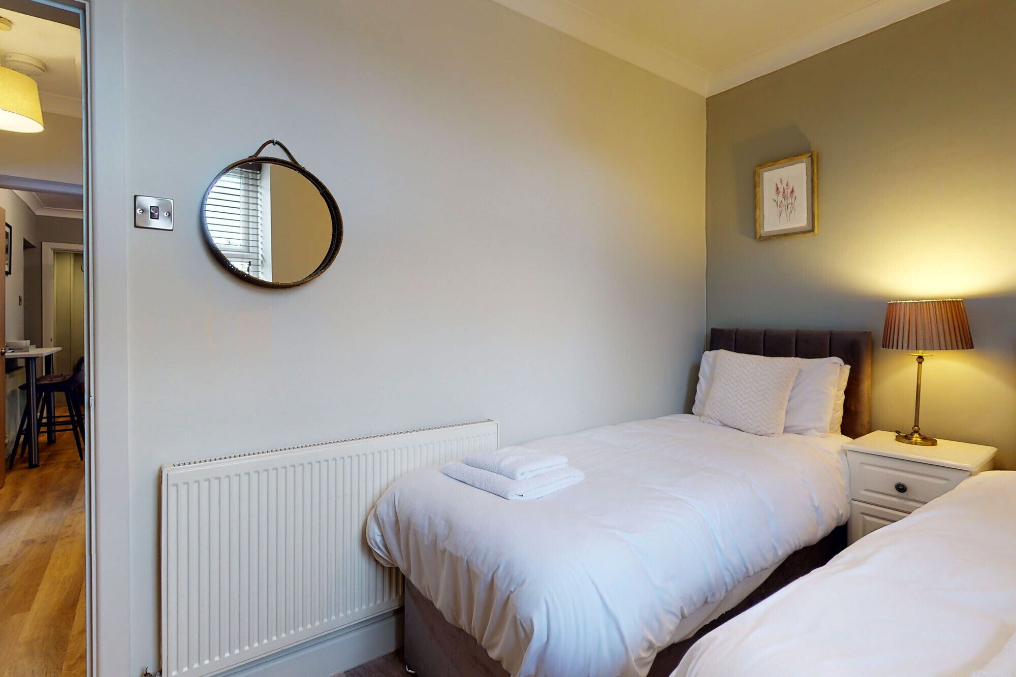 A hotel room with two beds, a round mirror, a radiator, a bedside table with a lamp, and a framed picture on the wall.