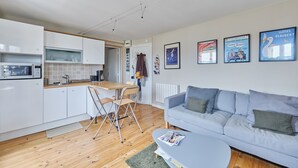 Cocina moderna con armarios blancos, encimera de madera y zona de comedor con mesa y sillas. Hay un sofá gris con cojines en la sala de estar y una revista sobre la mesa de centro.