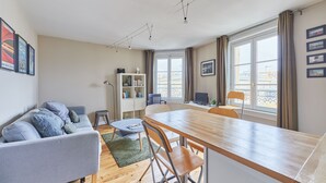 Una sala de estar moderna con sofá, mesa de comedor y sillas. Hay una estantería, un escritorio con computadora y una ventana con cortinas.