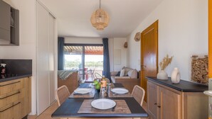 Un coin repas avec une table dressée pour deux personnes, une cuisine dotée de meubles en bois et une vue sur un espace extérieur aménagé, accessible par une porte vitrée.