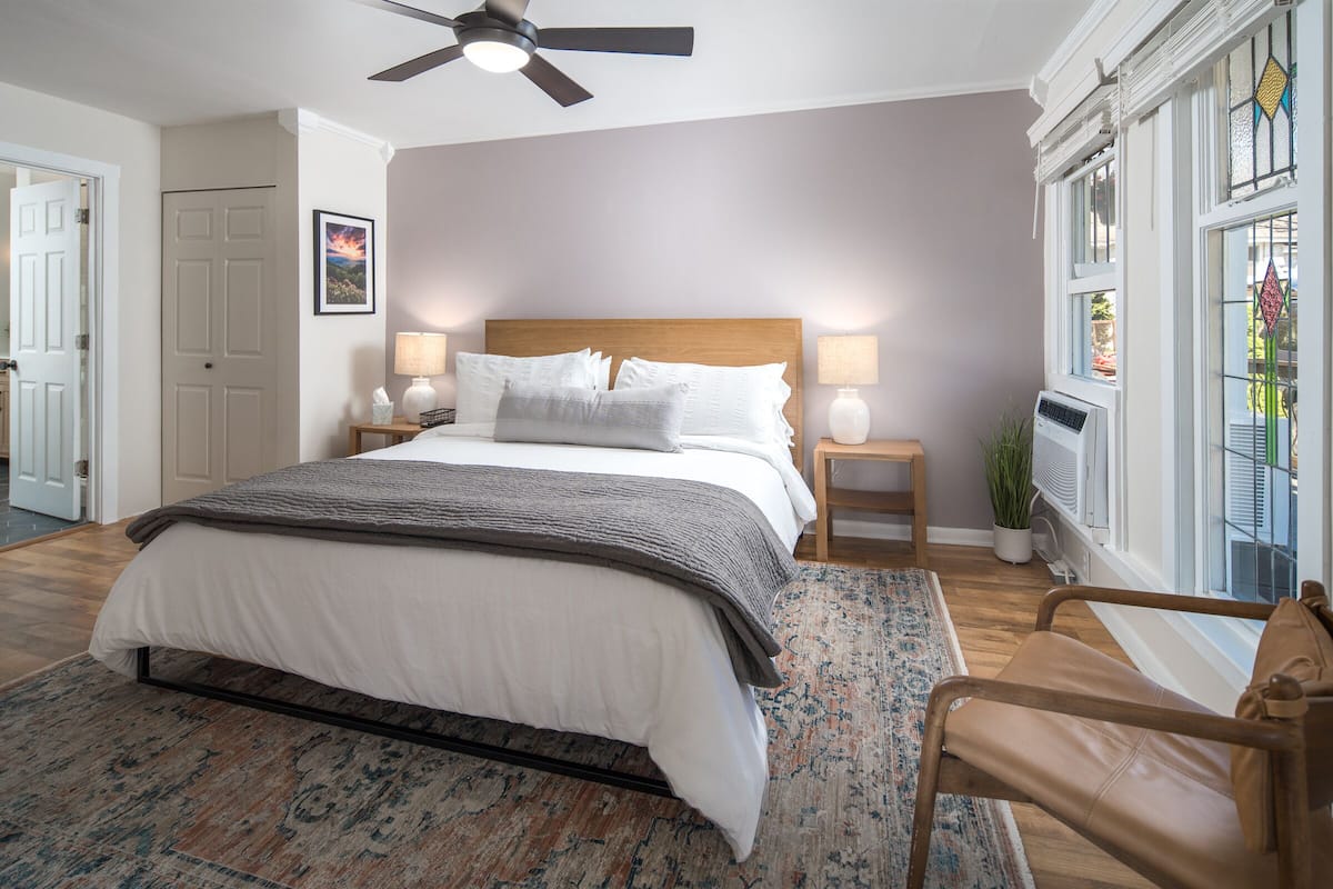 A bedroom with a large bed, a ceiling fan, a nightstand with a lamp, a window with a view, and a chair.