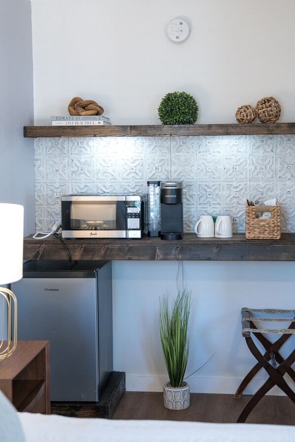 Una kitchenette compacta con microondas, cafetera y un refrigerador pequeño. En la repisa hay objetos decorativos, como una planta en maceta y una cesta tejida.